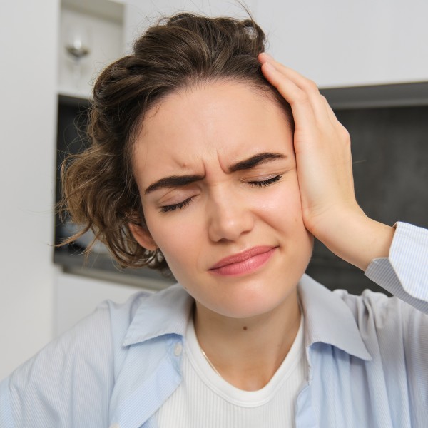 Nahaufnahme einer brünetten Frau, die sich den Kopf berührt, sich schwindelig fühlt, Migräne oder Kopfschmerzen hat und vor Schmerz das Gesicht verzieht.